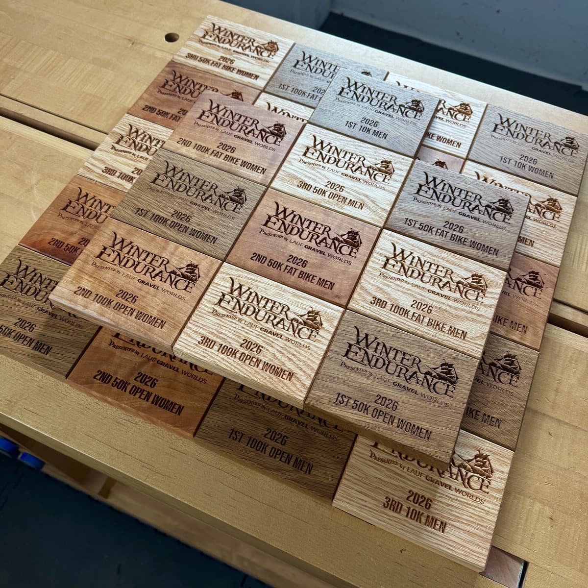Overhead view of complete set of Winter Endurance 2026 trophy plaques arranged in rows showing all wood species variations—white oak, red oak, and cherry—with CNC-engraved text for placement categories and race information