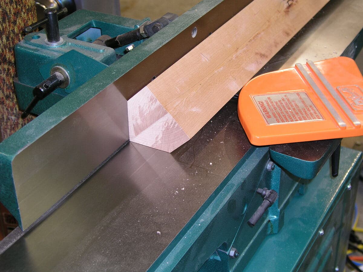Curved cherry leg in router jig setup on green jointer workbench with orange clamp and wood samples for finish testing