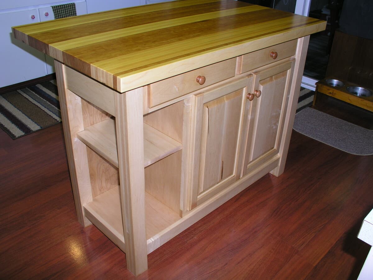 Kitchen island detail showing wooden drawer pulls, cabinet doors with raised panels, and open storage shelf with wood grain visible in natural light