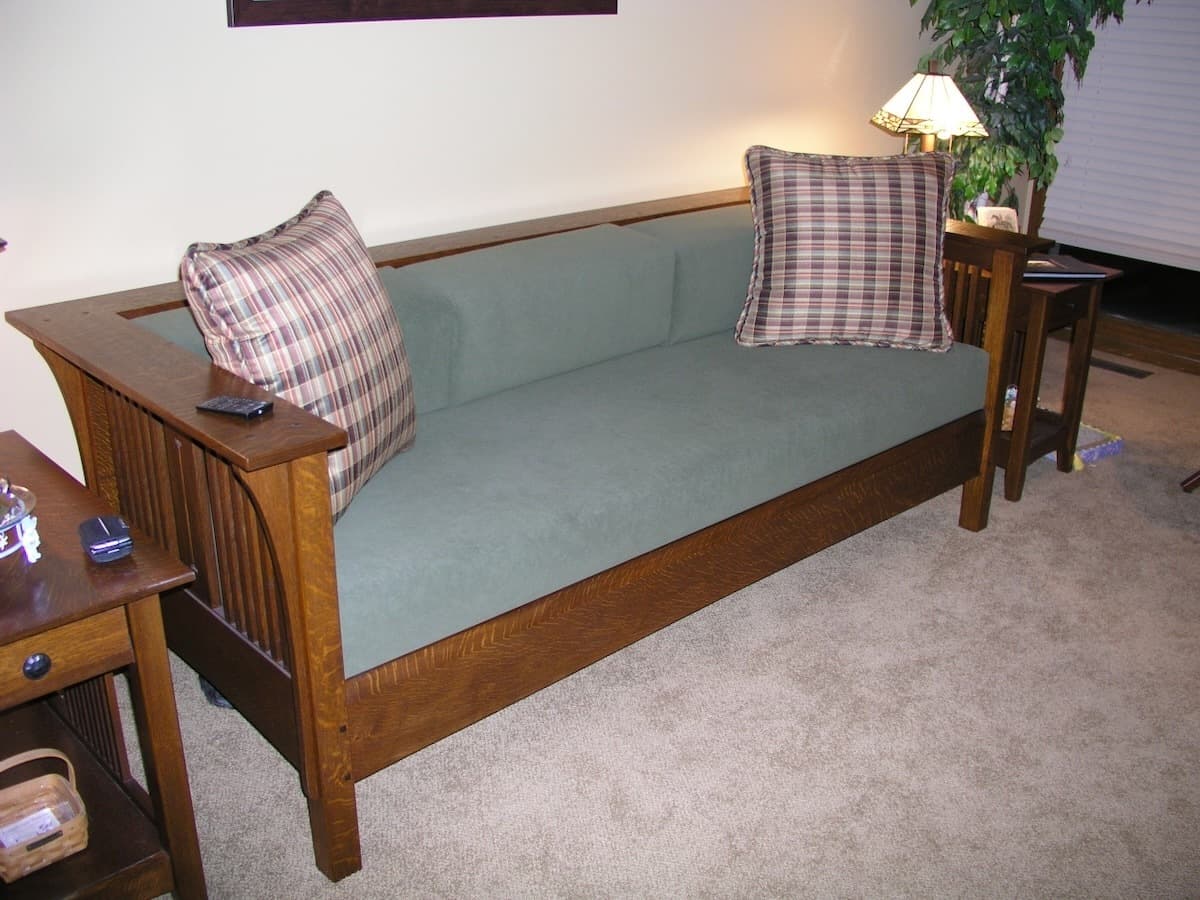 Completed Mission-style sofa in a living room setting with sage green cushions and plaid throw pillows. The quarter-sawn white oak frame features characteristic Mission-style vertical slat sides and arms, finished with a warm Shellac and Waterlox Satin that highlights the wood grain. The sofa is photographed in place with matching end tables, demonstrating its functional use as furniture.