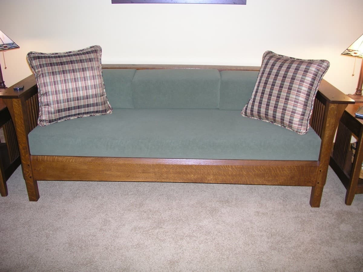 Front view of the completed Mission sofa showing the clean lines and symmetry of the design. The vertical slat backrest and side panels showcase the quarter-sawn white oak construction, while the sage green upholstered cushions contrast beautifully with the warm wood finish. Two plaid pillows complete the traditional Mission aesthetic.