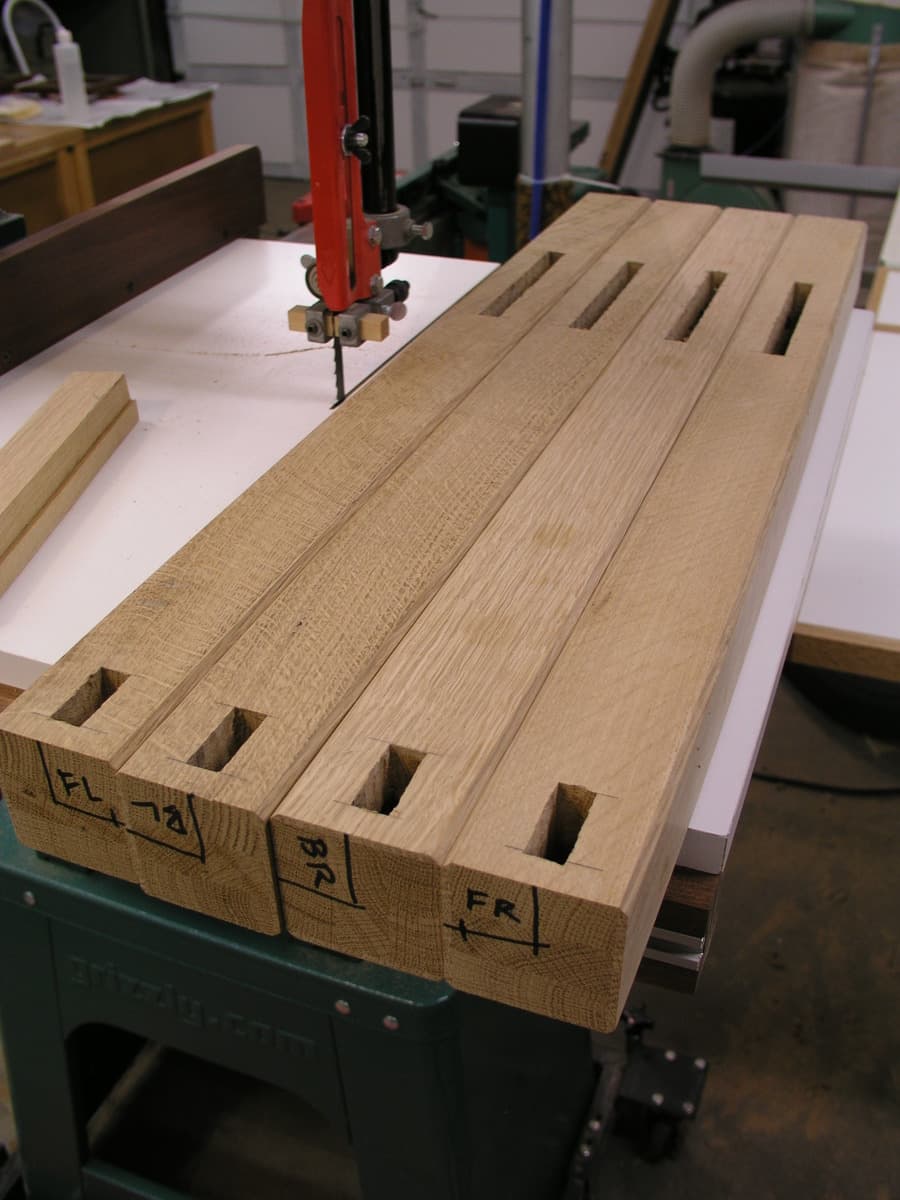Quarter-sawn white oak arm component during construction with mortise slots being drilled. The piece is mounted in a red drill press, showing precision mortises cut for the vertical slats of the Mission-style design. Pencil markings on the end grain indicate careful layout work, and the distinctive ray fleck pattern of quarter-sawn oak is visible.