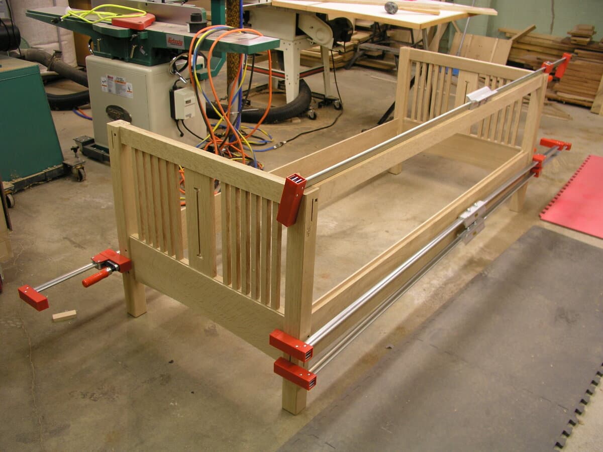 Full sofa frame assembly in progress, held together with red bar clamps during glue-up. The raw white oak frame shows both arms assembled with vertical slats, rails, and stretchers in place. This critical assembly stage ensures all mortise and tenon joints are properly aligned before the glue sets.