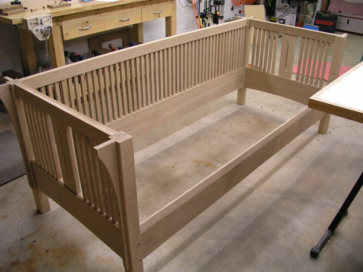 Detail view showing two walnut plugs flush-installed in quarter-sawn white oak arm component. The beautiful ray fleck pattern of the quarter-sawn oak is prominently displayed, with the dark walnut accents providing traditional Mission-style decorative detail. The wood appears unfinished or in early finishing stages.
