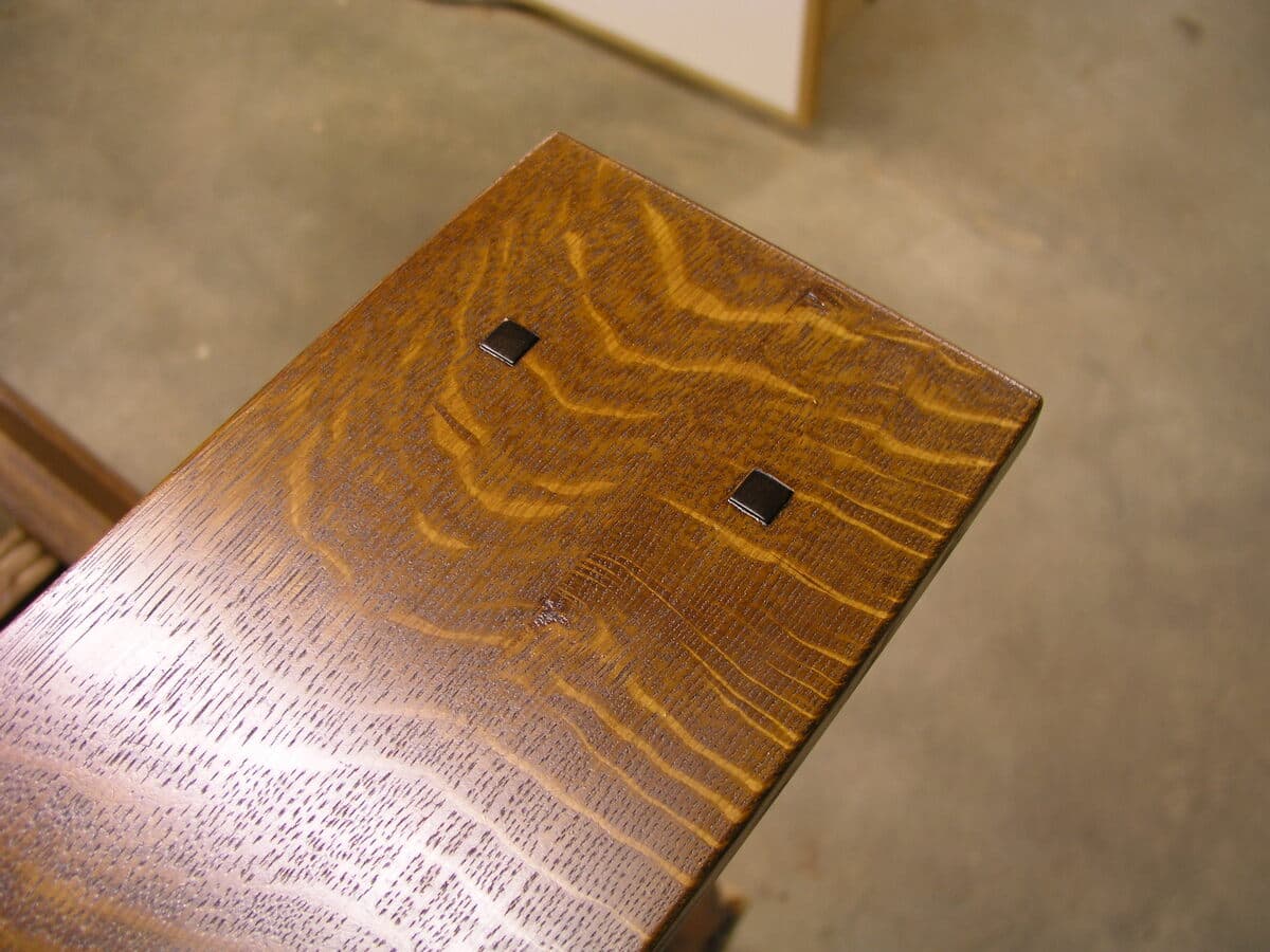 Overhead detail showing the finished arm with dramatic quarter-sawn oak figure highlighted by Shellac and Waterlox finish. The warm golden-amber color and prominent ray fleck pattern demonstrate premium lumber selection, with two walnut plugs visible as decorative accents. The vertical slat construction is visible in the background.