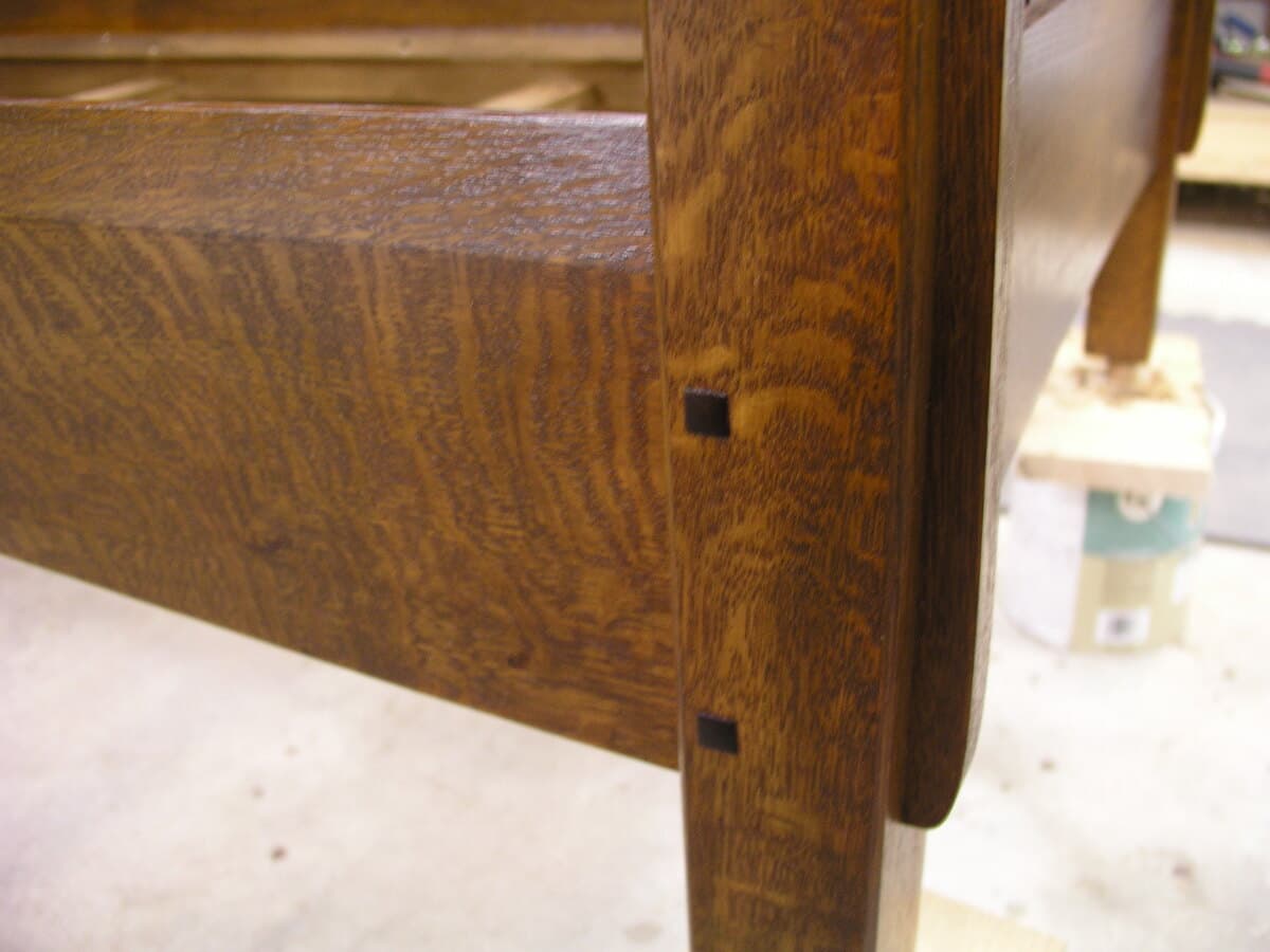 Detail of finished sofa arm showing mortise and tenon joint with walnut plug accent. The Shellac and Waterlox Satin finish brings out the warm honey tones and distinctive ray fleck of quarter-sawn white oak. The precise joinery and contrasting walnut plug exemplify traditional Mission furniture craftsmanship from 2006.