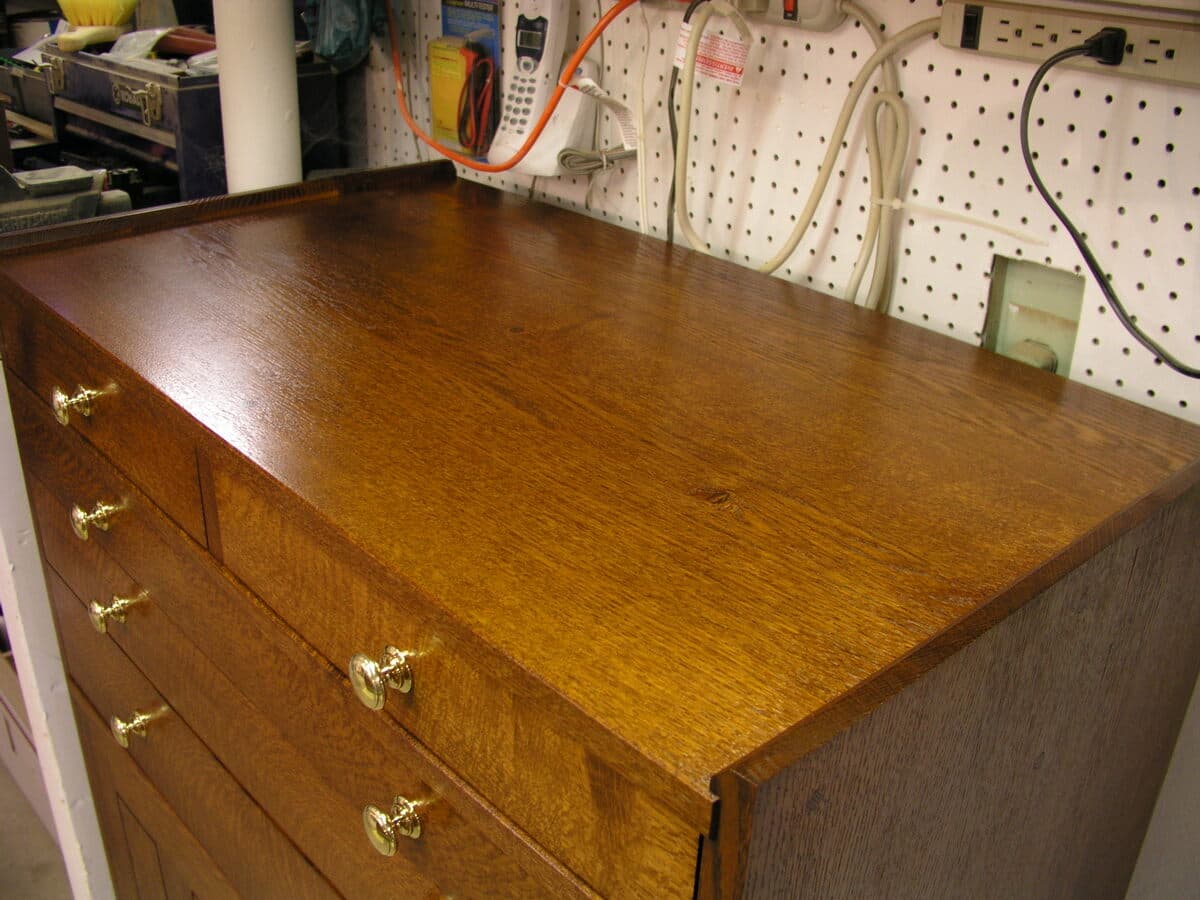 Close-up angled view of the tool cabinet's top surface showing the rich honey-brown rift-sawn red oak grain and the warm depth of the Shellac and Waterlox Satin finish. Brass pulls and the precise edge banding are clearly visible.