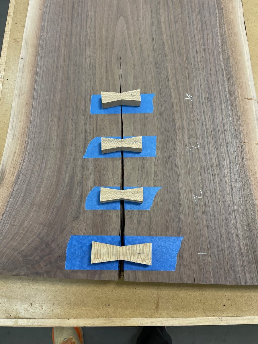 Walnut tabletop with four hand-cut sycamore bowtie inlays positioned along center crack, blue painter's tape marking router mortise locations during fabrication process.