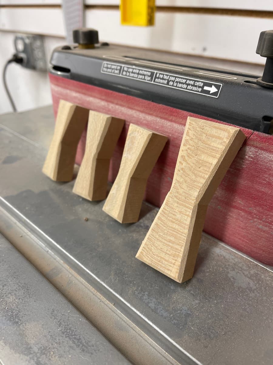 Four shaped sycamore bowtie blanks being sanded on oscillating edge sander, showing process of refining hand-cut bowties before final installation into walnut tabletop.
