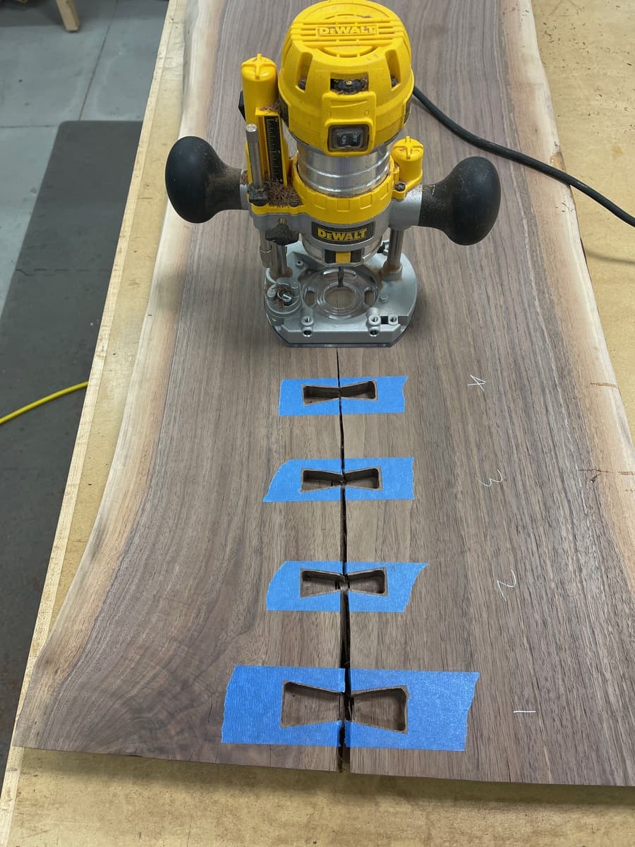 DeWalt plunge router positioned on walnut slab with blue-taped bowtie mortise templates, showing routing process for four inlay cavities along natural wood crack.