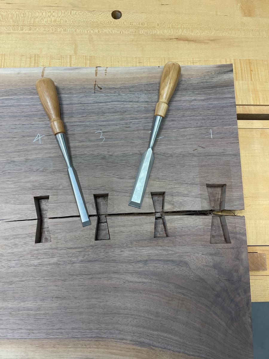 Two wood chisels resting on walnut slab showing four routed bowtie mortises along crack, demonstrating hand-chiseling process for precise fitting of sycamore inlays.