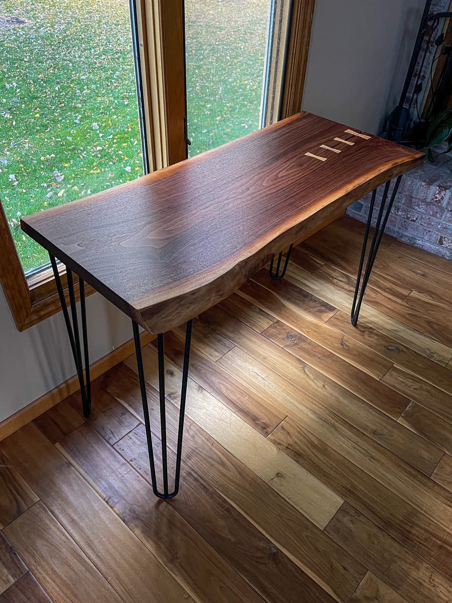 Finished walnut console table with black hairpin legs positioned by window, showcasing live-edge slab top with four sycamore bowtie inlays stabilizing center crack.