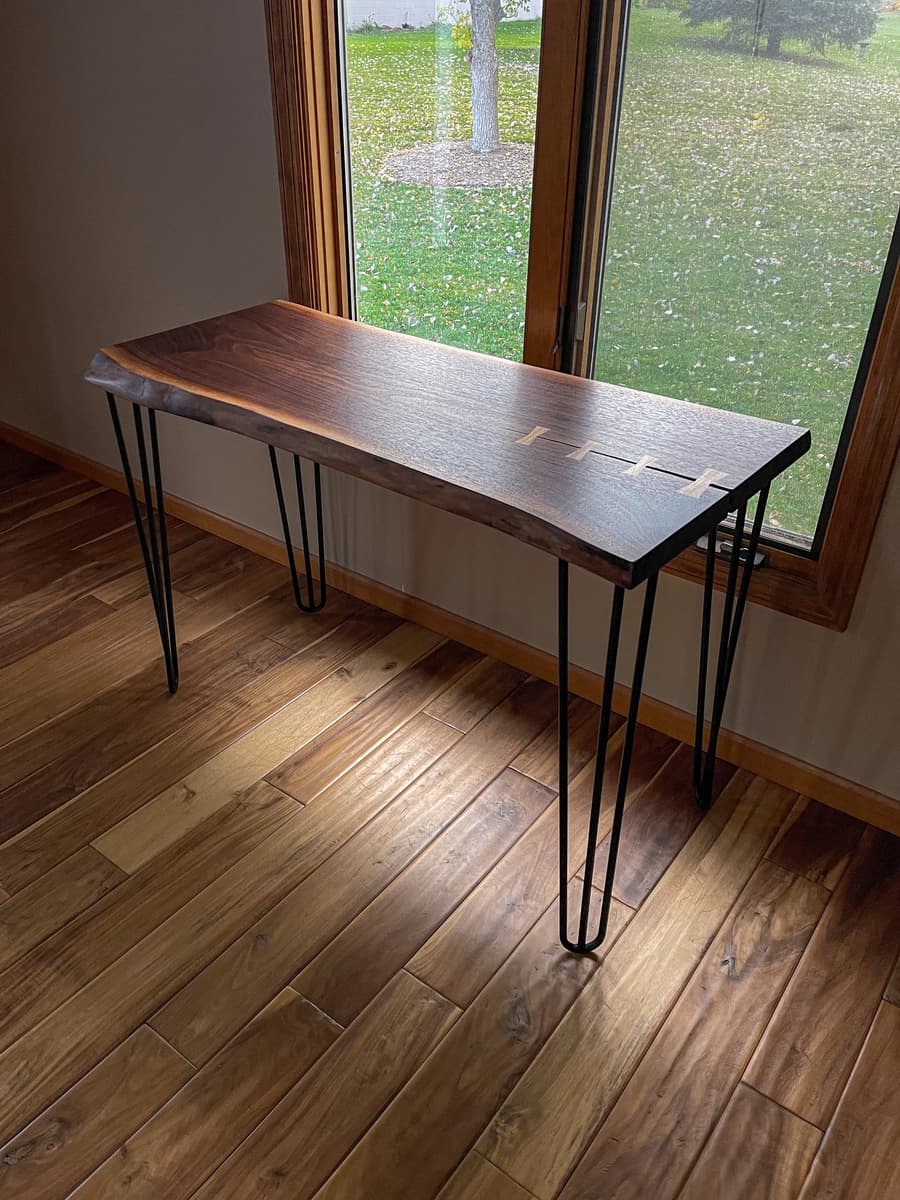 Full view of walnut console table with black hairpin legs by window, displaying natural live edge, rich grain patterns, and visible sycamore bowtie repairs.