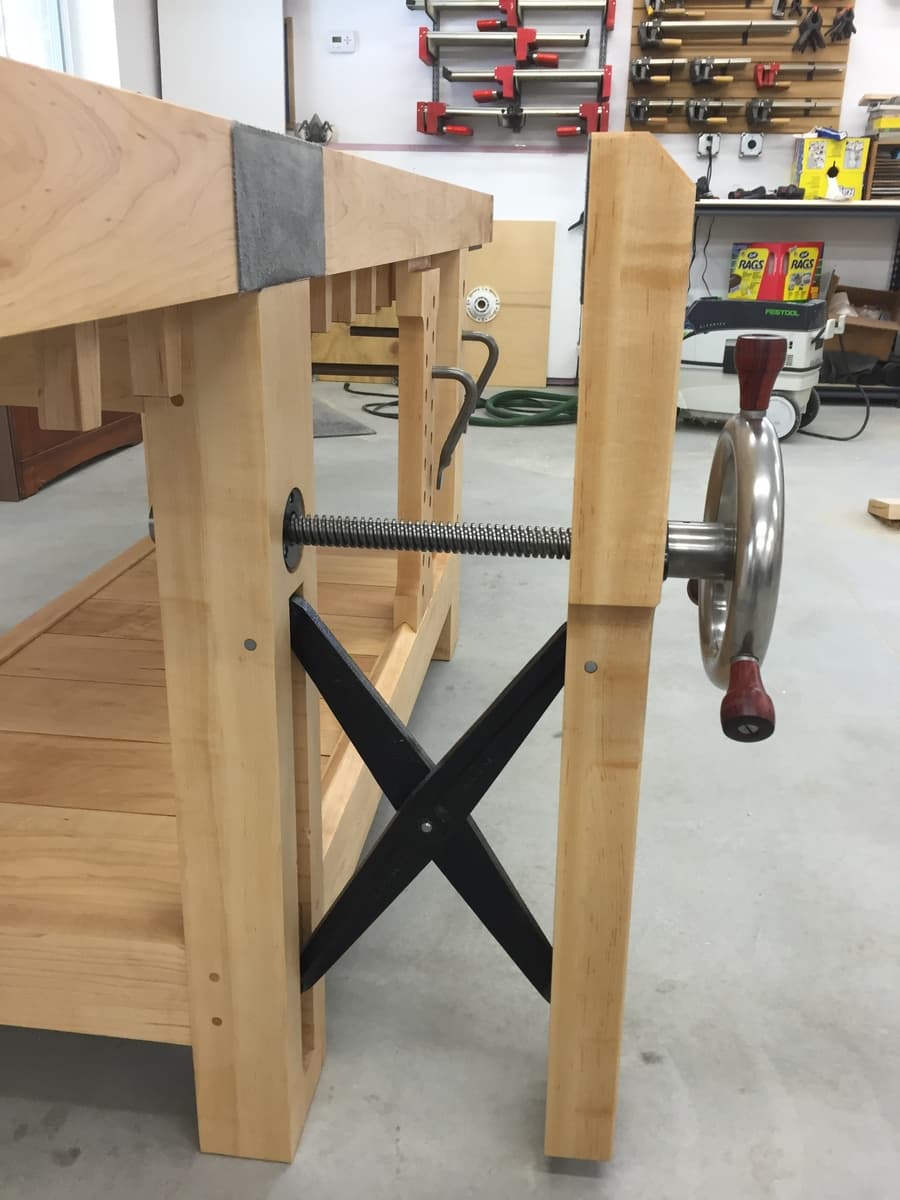 Completed hard maple Roubo workbench with leg vise featuring steel handwheel and black X-stretcher base, tail vise visible on opposite end