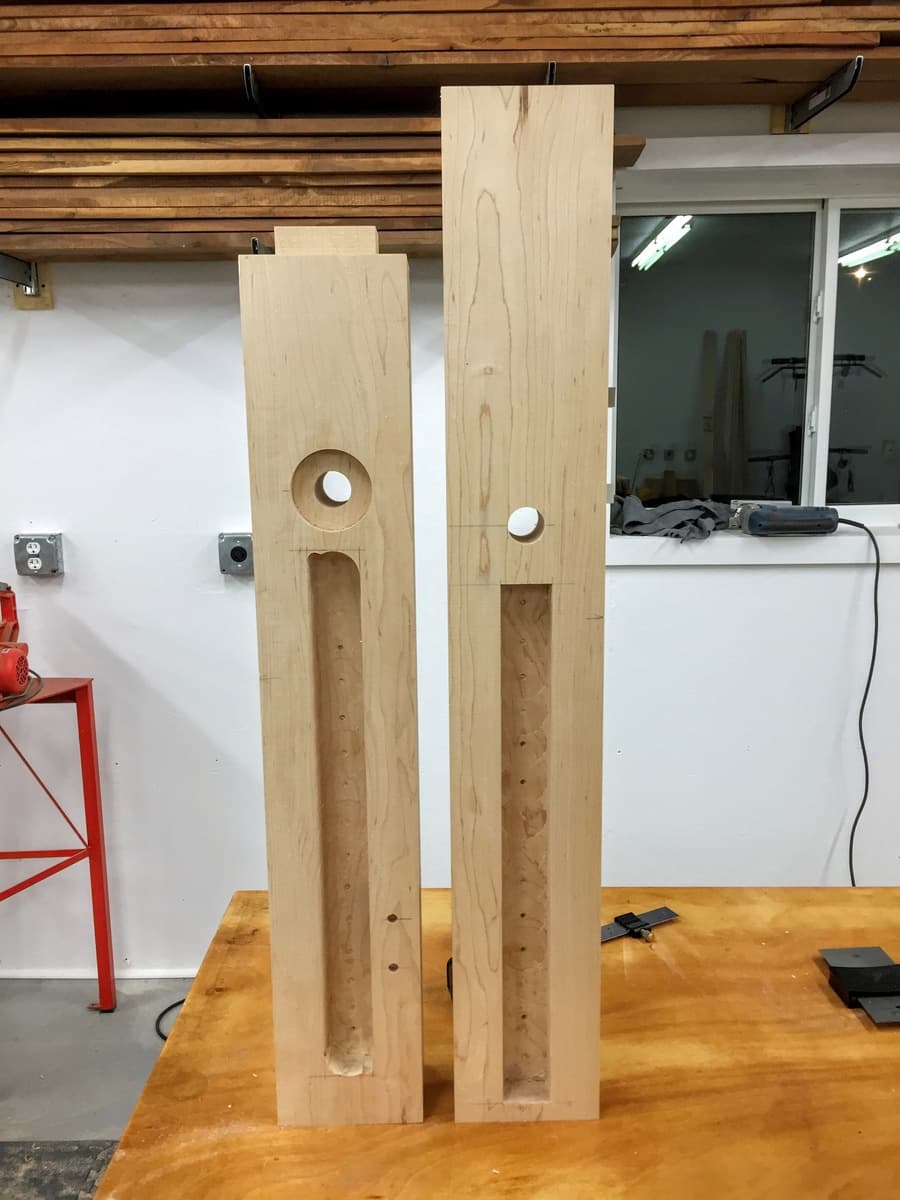 Two hard maple leg blanks standing upright showing circular and rectangular mortises for vise hardware and stretcher joinery