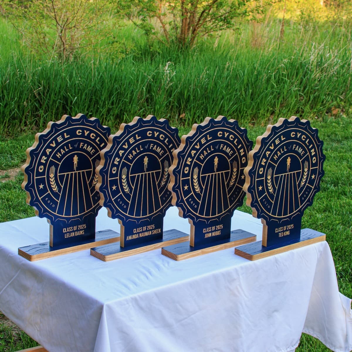 Four Gravel Cycling Hall of Fame trophy plaques displayed on white table with green grass background. Each features navy blue ash wood base with custom gold-engraved design for 2025 winners: Lelan Dains, Amanda Nauman Sheek, John Hobbs, and Ted King.